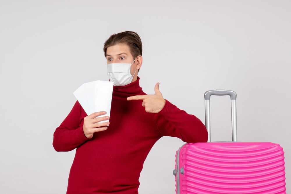 Front view of young male in mask holding plane tickets on a white wall