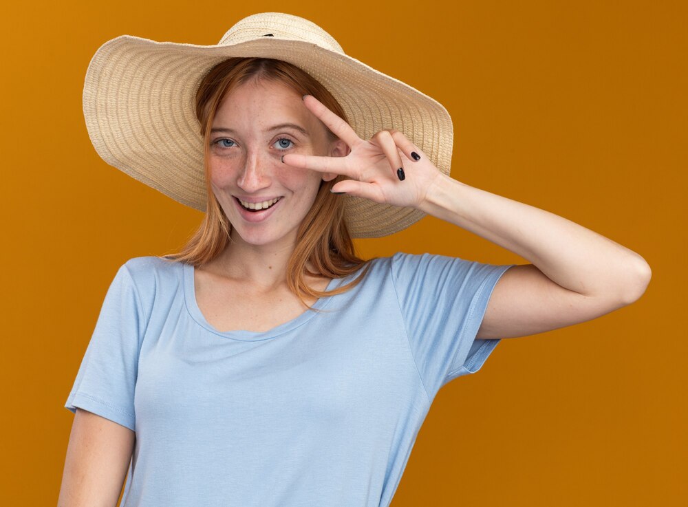 Pleased young redhead ginger girl with freckles wearing beach hat gesturing victory sign on orange